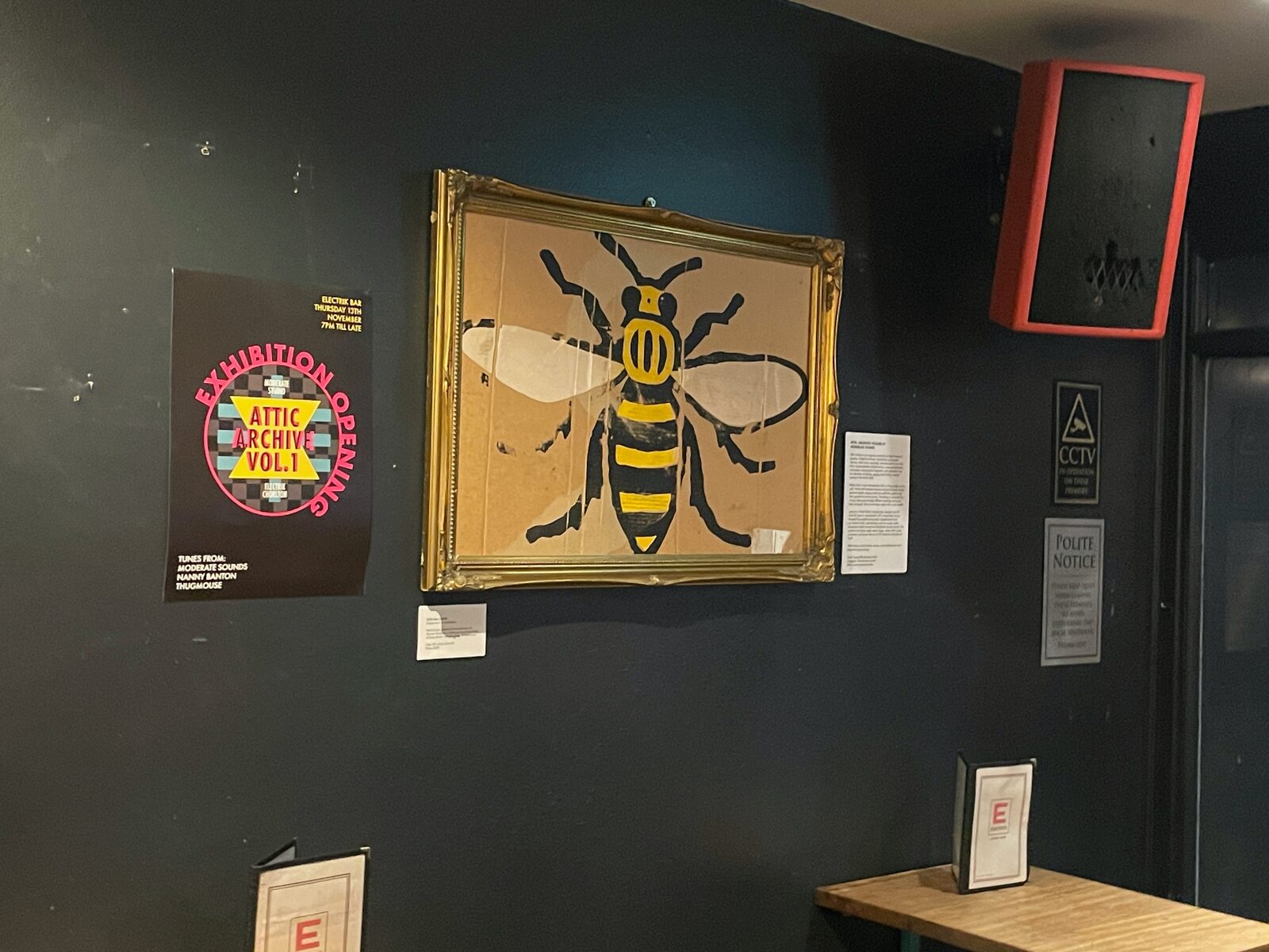 The Attic Archive Vol. 1 exhibition poster beside a framed worker-bee print at Electrik.