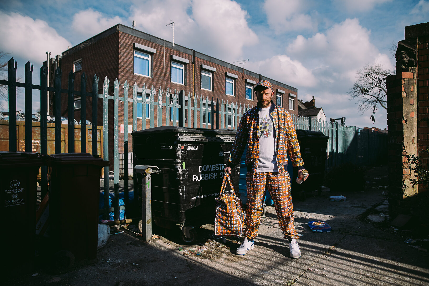Mark Jermyn wearing a custom tracksuit and matching bag made from a distorted Union Jack pattern, standing beside wheelie bins in Manchester