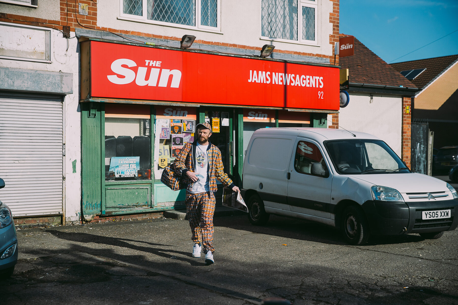 Mark Jermyn leaving a newsagent under a Sun newspaper sign, wearing the Brexit tracksuit and carrying the matching bag and a copy of The Sun