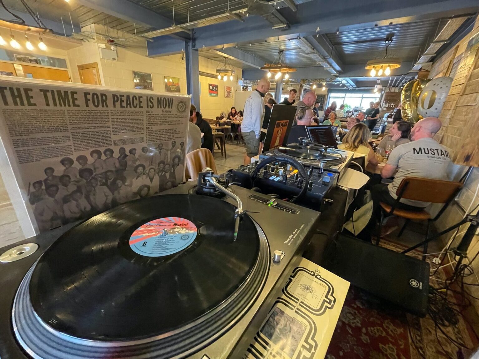 A record playing on a turntable in the foreground, with people sitting at tables in the room beyond