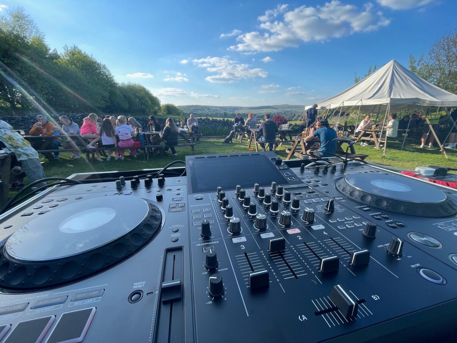 A DJ controller in the foreground looking out over a field of picnic tables and a marquee under a blue sky