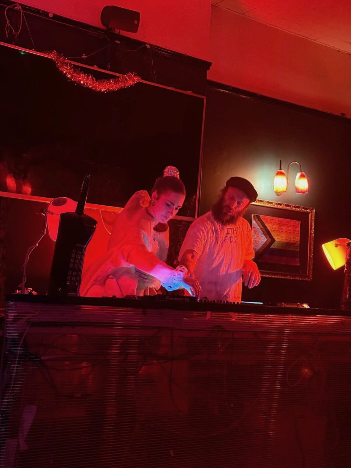 Two DJs playing side by side behind the decks in a room lit deep red