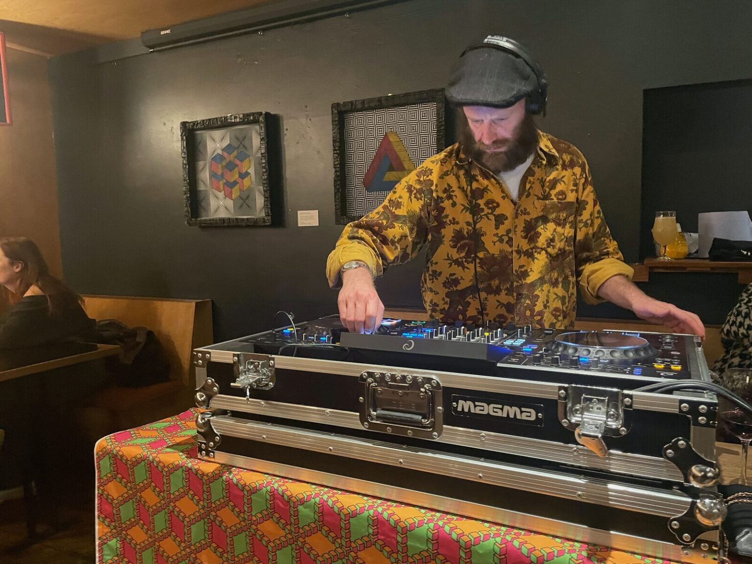 A DJ in headphones playing at a controller set on a flight case in a bar, framed pictures on the wall behind