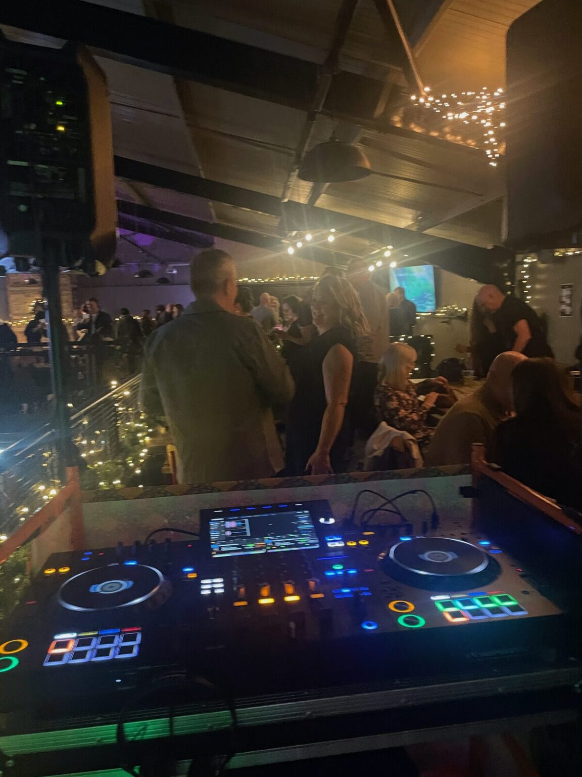 A lit DJ controller in the foreground with people standing and talking in the room beyond
