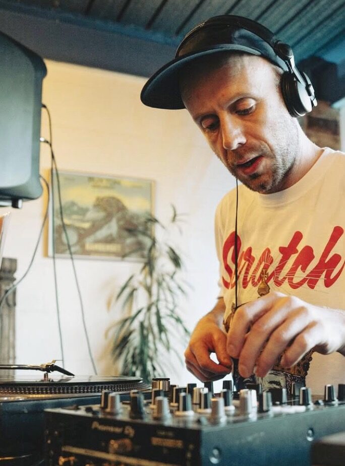 A DJ in headphones leaning over a mixer and turntable, with a plant and a framed picture behind