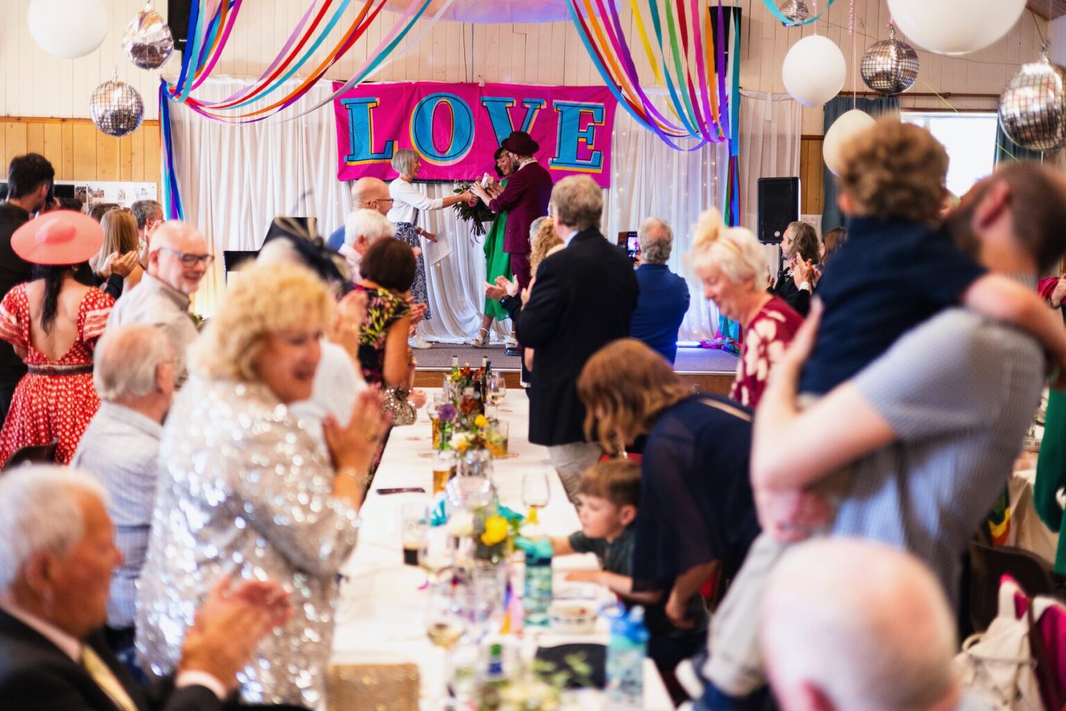 Guests along a long table, cheering towards a stage hung with a LOVE sign, ribbons and mirrorballs overhead