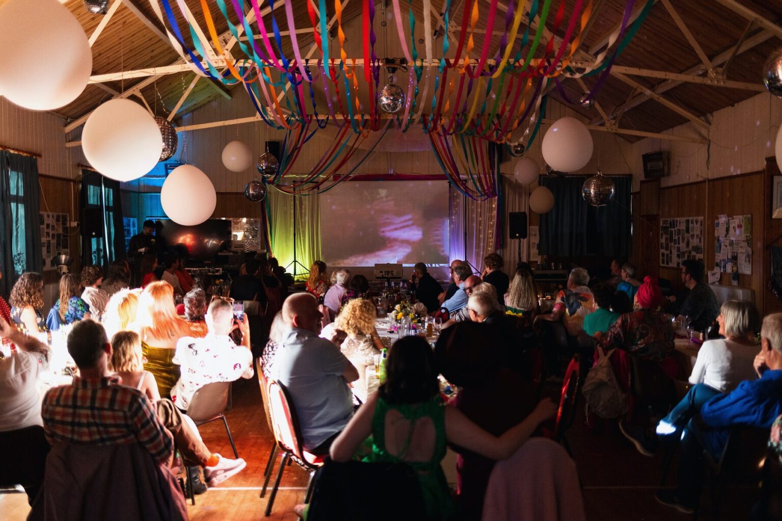 A hall set for a party, coloured ribbons and balloons strung across the beamed ceiling above seated guests