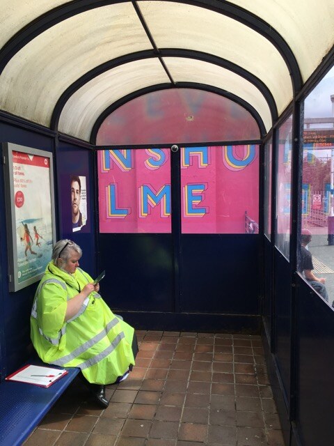 A station worker sitting in the platform shelter with the Levenshulme Station Mural visible through the windows