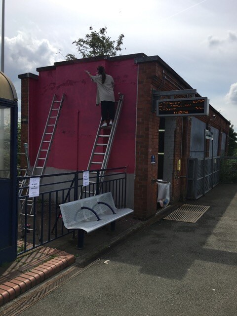 Mark Jermyn painting the pink ground coat of the Levenshulme Station Mural from a ladder