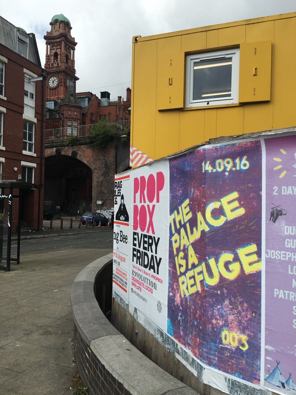 A hand screen-printed poster reading The Palace Is A Refuge, wheatpasted onto a Manchester street corner with the Palace Hotel clock tower visible behind