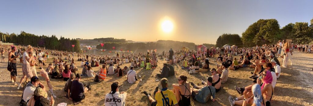 A wide view of a crowd sitting on dry grass at Boomtown in low evening sun, dust hanging in the air