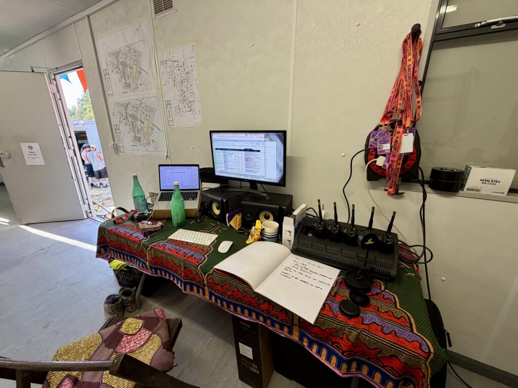 The photography office at Boomtown, a desk with the day schedule up on the monitor, radios charging and site maps pinned to the wall